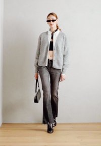 Cardigan in maglia grigio chiaro sopra un top corto nero, abbinato a jeans grigi a zampa e stivaletti neri con tacco. Gli accessori includono occhiali da sole e una borsa piccola.