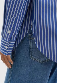 Blau-weiß gestreiftes Hemd mit geknöpften Manschetten, das über einem Jeansrock hängt. Sichtbare Tasche und kontrastierende Nähte am Stoff.