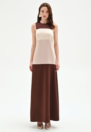 Femme debout portant une robe maxi sans manches à empiècements de couleur marron, beige et crème, avec des boucles d'oreilles dorées et des sandales métalliques.