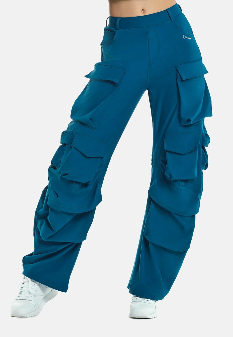 Pantaloni cargo verde acqua con molte tasche ampie, vestibilità comoda, texture arricciata lungo le gambe e vita aderente; abbinati a sneakers bianche.
