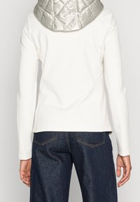 Bílá bunda s dlouhými rukávy a lesklou prošívanou stříbrnou kapucí; doplněná tmavými džíny. Čisté linie a jednoduchý design.