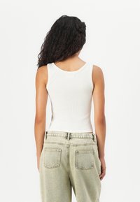 Woman with long curly hair wearing a white sleeveless ribbed top and light-washed high-waisted jeans, shown from the back against a white background.