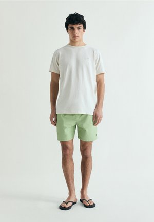 Joven de pie, vestido con una camiseta blanca lisa, pantalones cortos verde claro y chanclas negras, sobre un fondo gris claro.