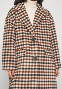Braun-, Schwarz- und Cremefarbener Karo-Mantel aus einem weichen Stoff mit großem Kragen, zwei Fronttaschen und schwarzen Knöpfen auf der Vorderseite.