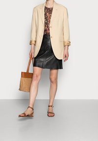 Person som bär en beige kavaj över en blommig topp, svart läderskjort, bruna ankelremssandaler och håller en brun vävd tote-väska.