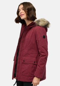 Bordo parka su dirbtiniu kailiu, zip ir spaudžių uždarymu, šoninėmis kišenėmis ir šiek tiek tekstūruotu audiniu. Sukurtas su insignija ant rankovės.