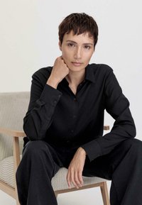 Black button-up shirt in a soft fabric with a collar; model seated on a textured light-colored chair, one hand resting on chin.