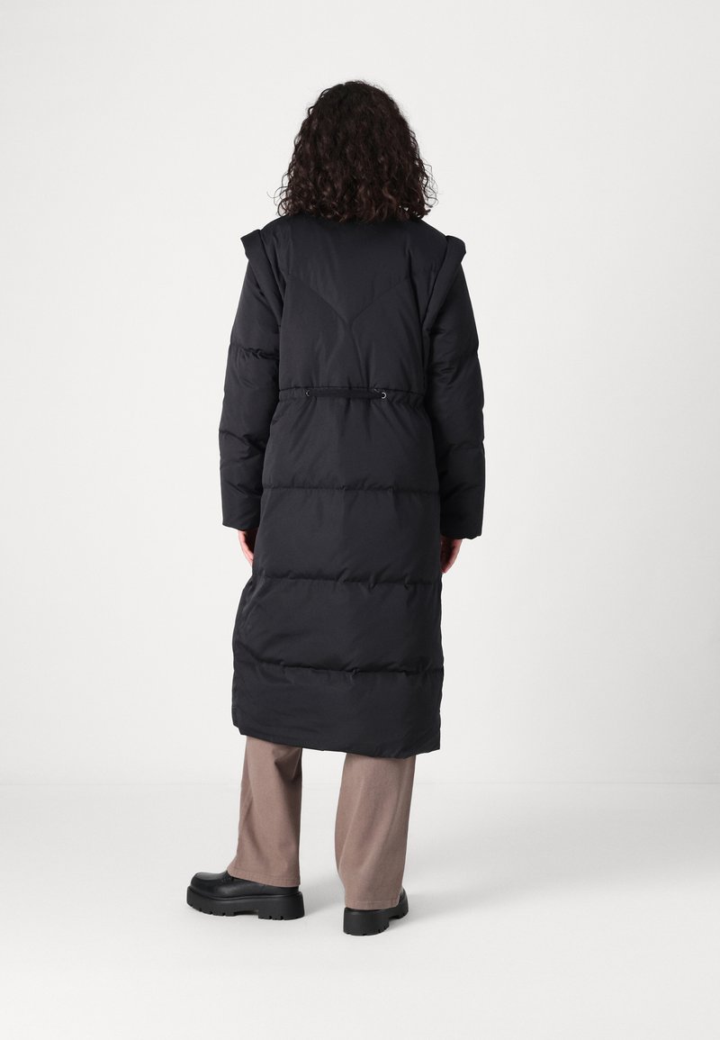 Schwarze gepolsterte Jacke mit langem Design, ausgestattet mit einer Kordelzug-Taille, breiten Schulterdetails und einer glatten Textur. Getragen über beigefarbenen Hosen und schwarzen Schuhen.