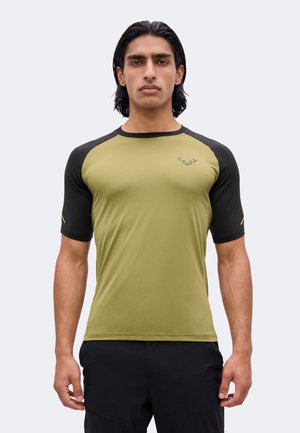 Uomo con capelli neri di media lunghezza che indossa una maglietta sportiva a maniche corte verde oliva e nera, in piedi davanti a uno sfondo chiaro e uniforme.