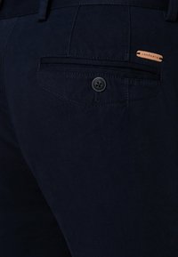 Zbliżenie materiałowych spodni w kolorze granatowym, z teksturowaną powierzchnią, tylną kieszenią z guzikiem oraz skórzaną metką z logo przyszytą nad kieszenią.