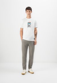 Camiseta blanca con personaje de dibujos animados y el texto "GENIUS", combinada con pantalones gris claro y zapatillas beige. Look sencillo y casual.