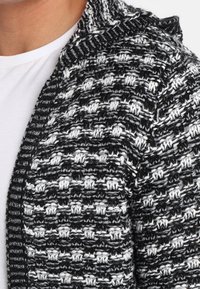 Cardigan lavorato a maglia in bianco e nero, con motivo a trama, apertura frontale e collo ampio. Il tessuto è morbido e leggermente spesso.