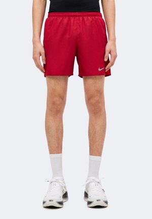Osoba w czerwonych spodenkach sportowych, białych skarpetkach sportowych i białych butach do biegania Nike, stojąca na jednolitym tle.