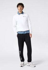 Sudadera blanca con logo, superpuesta sobre una camisa de mezclilla. Pantalones deportivos negros con logo blanco, combinados con zapatillas deportivas grises. Textura suave.