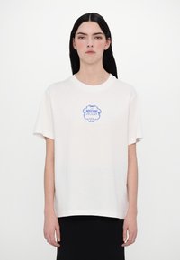 Balts kokvilnas T-kreklis ar zilu grafisko drukājumu, kas lasāms "MOSCHINO JEANS HOTEL". Kreklam ir īsas piedurknes un brīva piegriezuma.