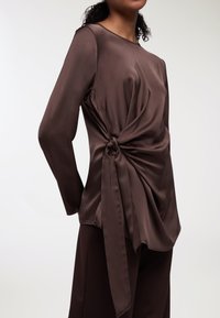 Top lungo a maniche con nodo laterale in satin marrone, caratterizzato da un collo rotondo e una superficie liscia. Abbinato a pantaloni scuri coordinati.