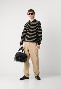 Pullover rayé noir et olive, pantalon beige, lunettes de soleil noires, tenant un sac en cuir noir et gris avec des bords arrondis et des poignées courtes.
