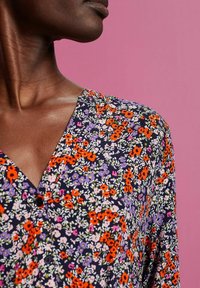 Lähikuva henkilöstä, joka on pukeutunut kukkamekkoon, jossa on oransseja, purppuran, vaaleanpunaisia ja valkoisia kukkia tummalla taustalla pinkkiä taustaa vasten.