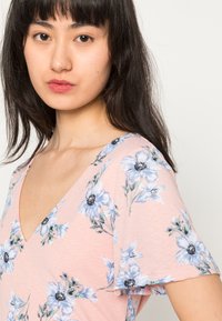 Top rosa floral de manga curta com um decote em V profundo. Apresenta flores azuis e folhas verdes em um tecido suave e texturizado.