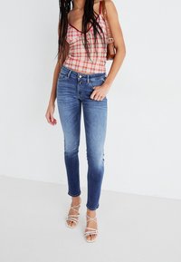 Jeans pitillos azul claro con un acabado suave, combinados con una blusa de cuadros rojos y blancos. La modelo lleva tacones blancos con tiras y un bolso marrón.