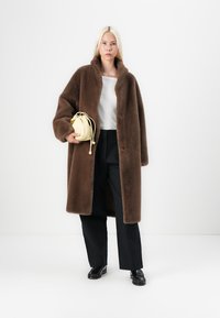 Manteau en fourrure synthétique marron avec un col, boutons uniques, associé à un top blanc et un pantalon noir à jambes larges. Tenant un petit sac jaune.