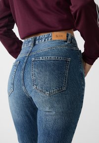 Högmidjade blå denimjeans med en blekt textur, distinkta bakfickor och en beige läderetikett sydd på midjebandet.