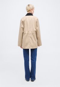 Άτομο με κοντά ξανθά μαλλιά που φοράει μπεζ σακάκι με μαύρο γιακά και μπλε τζιν, στέκεται γυρισμένο προς τα πίσω μπροστά σε φωτεινό φόντο.