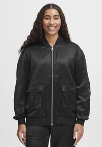 Jachetă bomber din satin negru, cu fermoar frontal, două buzunare frontale cu clapetă, manșete ribbate și talie ajustată. Textură netedă cu un design elegant.