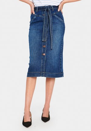 Dżinsowa spódnica midi z guzikami z przodu i związanym paskiem w talii, noszona z czarnymi szpilkami z ostrym noskiem i rękami na biodrach.