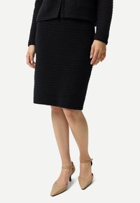 Kvinna som bär en svart texturerad knälång kjol och matchande långärmad topp, kombinerat med beiga ankelbandskor med spetsig tå.