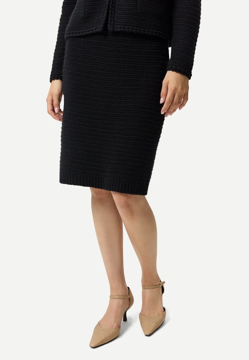 Kvinna som bär en svart texturerad knälång kjol och matchande långärmad topp, kombinerat med beiga ankelbandskor med spetsig tå.