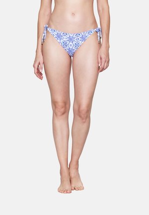 Frau trägt blaue und weiße Bikinihose mit Blumenmuster und seitlichen Knoten, steht aufrecht vor schlichtem hellem Hintergrund.