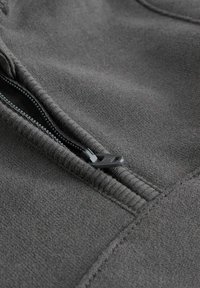Grauer Sweatshirtstoff mit strukturiertem Finish. Verfügt über einen schwarzen Reißverschluss und Nähte entlang der Säume, die das Design des Kleidungsstücks betonen.