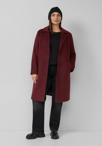 Burgunderfarbener Wollmischungs-Mantel mit Reverskragen, Knöpfen vorne und Taschen, kombiniert mit einem schwarzen Oberteil, schwarzen Hosen und glänzenden schwarzen Schuhen.