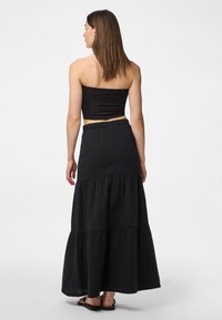 Femme aux cheveux bruns raides porte un haut court noir sans bretelles, une longue jupe noire à volants et des sandales plates noires, debout de dos.