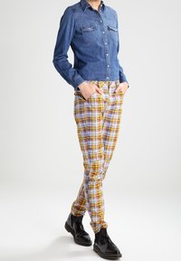 Jeansskjorta med två bröstfickor, blå färg; rutiga byxor i gula, rosa och lila nyanser; blanka svarta ankelstövlar.