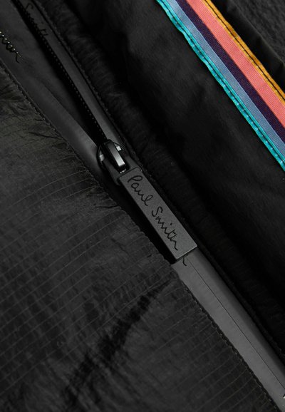 Melnā rāvējslēdzējs ar "Paul Smith" tekstu uz vilkšanas cilpas, piestiprināts pie teksturēta melna auduma ar daudzkrāsainu svītru apmalēm.