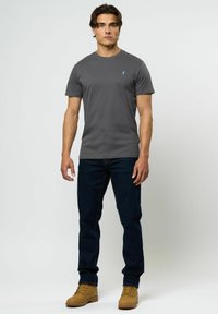 Grå t-skjorte med korte ermer og rund hals, kombinert med mørkeblå jeans og brune snøresko. Diskret logo på brystet.