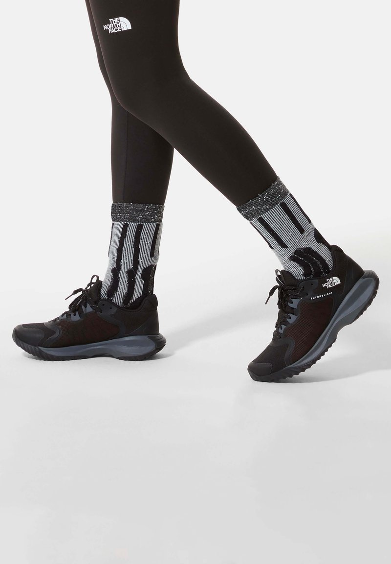 Nogi w czarnych legginsach The North Face i czarnych butach sportowych z szarymi podeszwami, noszone z czarno-białymi skarpetkami w wzory na białym tle.