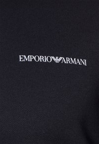 Černá žebrovaná látka s bílým logem "EMPORIO ARMANI" a stylizovaným akcentem orla, umístěným napříč ve středu.
