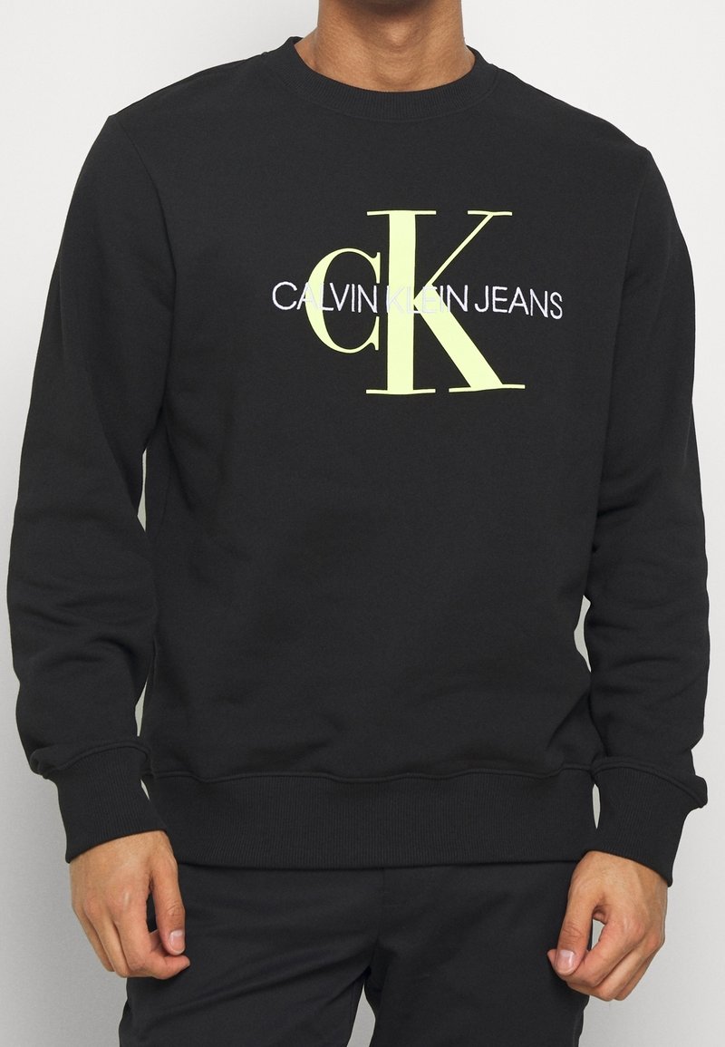 Černá mikina vyrobená z bavlny, s velkým žlutozeleným grafickým logem "CK" a textem "CALVIN KLEIN JEANS" v bílé barvě. Ribbené manžety a lem.