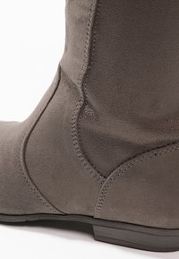 Stivale in suede grigio con una texture liscia, forma arrotondata e dettagli di cucitura visibili lungo il lato e la base. Design della suola bassa e piatta.