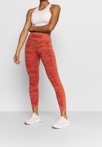 Leggings rossi a camo di Puma con una vita alta, tessuto testurizzato, dettagli nei pannelli laterali, abbinati a scarpe sportive rosa chiaro.