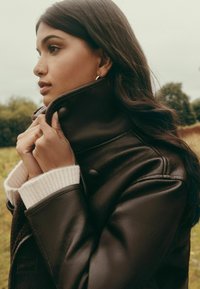 Pruun nahktagi kõrge kraega ja soonikkoes kreemjad varrukaotsad. Modell hoiab kätega kraed kinni, esitades jakikanga tekstuuri ja disaini.