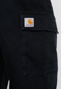 Zwarte cargobroek van een gestructureerde stof. Heeft een opvallend wit label met een oranje logo op een zijzak.