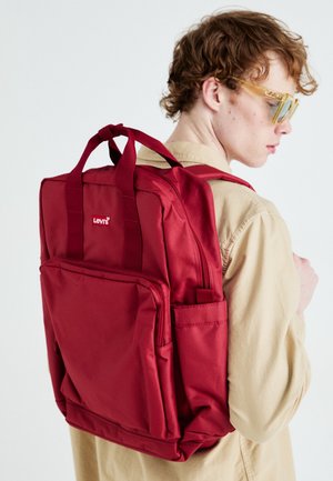Jeune personne portant une chemise beige et des lunettes de soleil jaunes porte un sac à dos rouge Levi's sur l'épaule sur fond blanc.