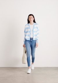 Cardigan rayé bleu et blanc avec des boutons, porté par-dessus un t-shirt blanc, associé à un jean bleu clair et des baskets blanches.