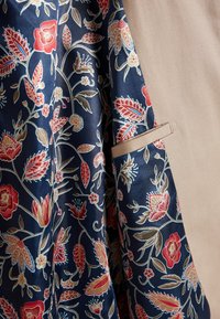 Blomstret stof i marineblå, med røde og cremefarvede blomster, kombineret med en beige tekstureret polyesterbaggrund og en skjult lomme.