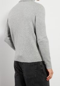 Personne portant un pull en laine gris clair et un jean noir, vue de dos avec le bras droit détendu.