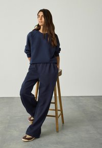 Frau mit langen Haaren, die einen navyblauen Sweatshirt und weite Hosen trägt, lehnt auf einem Holzschemel auf einem grauen Boden.